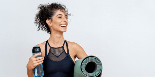 woman in active gear standing against a wall smiling and holding a water bottle and yoga mat