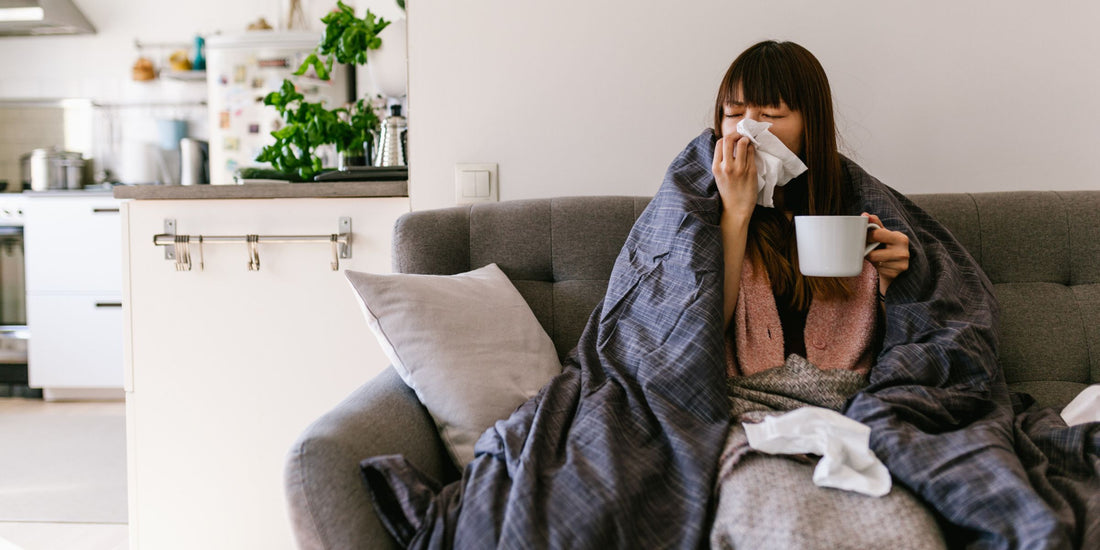 a person wrapped in a cozy blanket, cradling a warm mug, and sitting on a comfortable couch with tissues blowing their nose