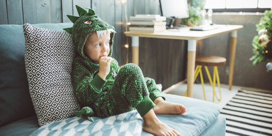 A child wearing a green, textured dinosaur costume is lounging on a grey sofa with a patterned cushion